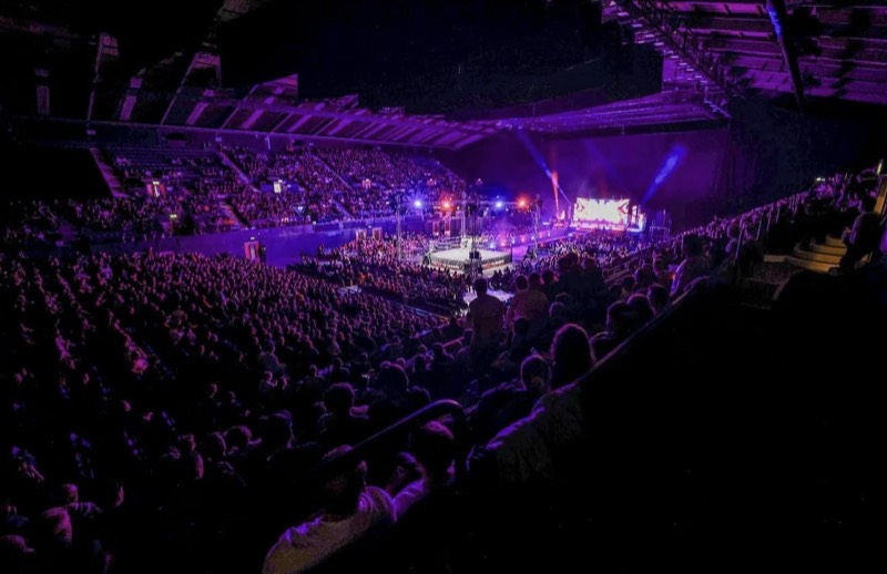 Packed arena around a lit wrestling ring with an LED screen