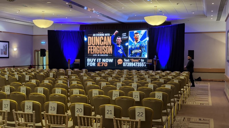 Function room set for a speaker evening with an LED screen, drape and blue uplighting